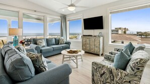 Un salon moderne avec un canapé, un fauteuil et une table basse, de grandes fenêtres offrant une vue sur la plage, et un téléviseur à écran plat fixé au mur.