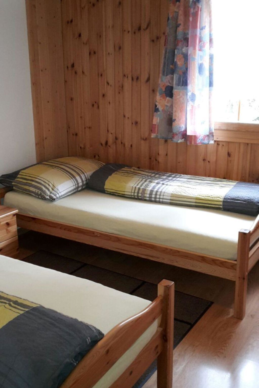 A wooden bunk bed with a mattress and pillows, a wooden nightstand, and a window with a floral curtain.