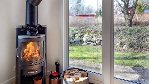 A wood-burning stove with a glass door, a metal pot, and a bucket of firewood next to a window with a view of a garden and a red building. - 3 bedroom beautiful home in Hörby (Hörby)