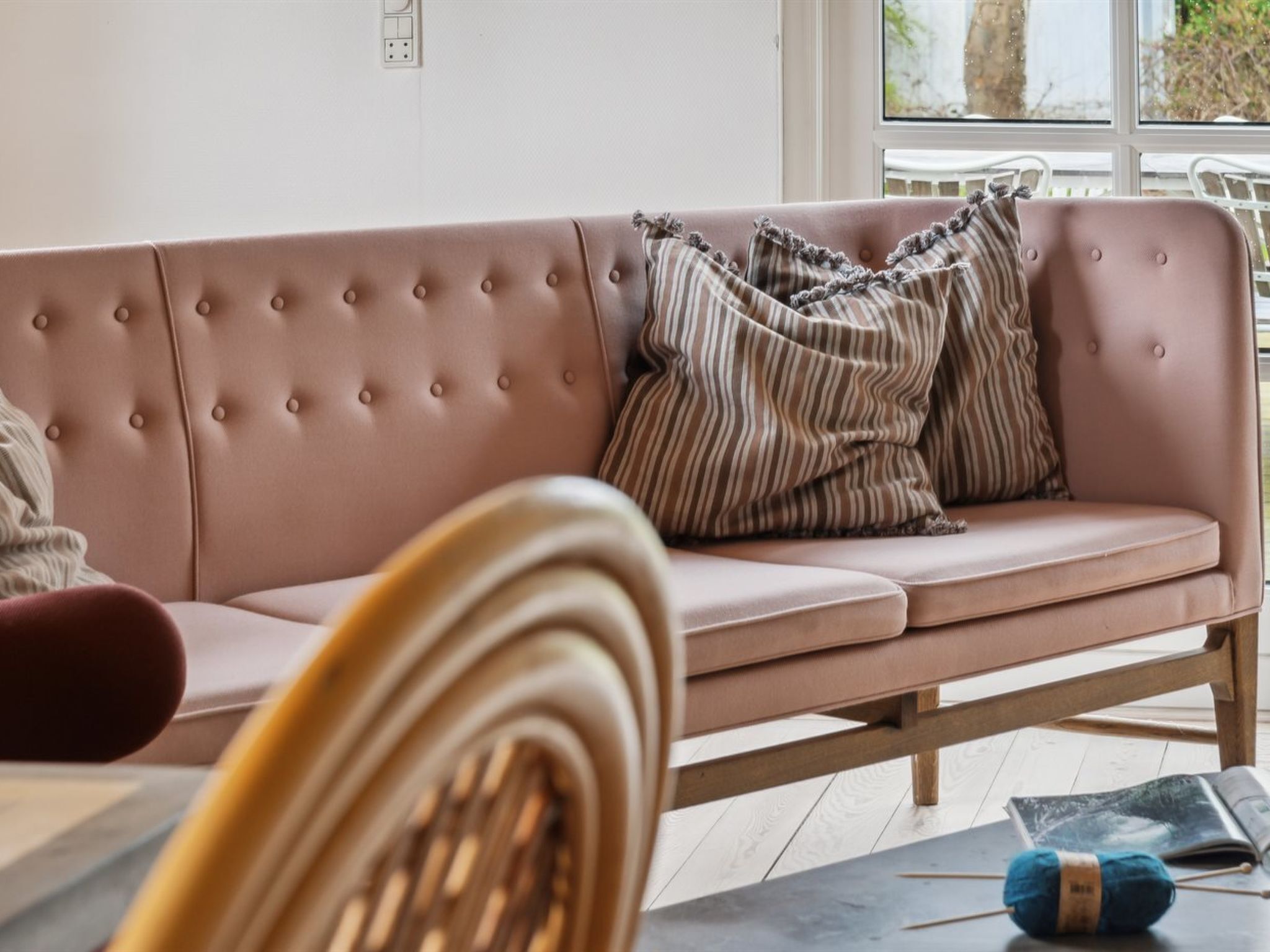 Un canapé rose à motifs capitonnés, agrémenté de coussins à rayures, une table en bois sur laquelle reposent un magazine et une pelote de laine bleue, et une fenêtre donnant sur des arbres.