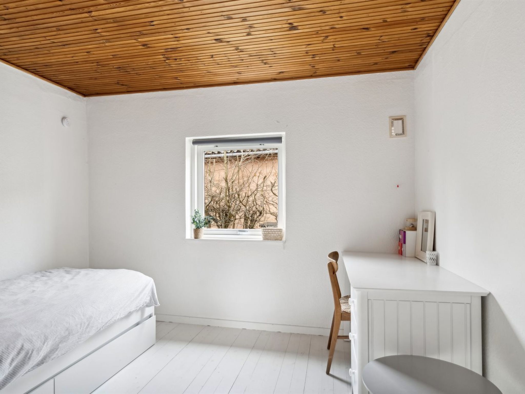 Une chambre avec un plafond en bois, une fenêtre donnant sur les arbres, un lit, une chaise et un petit bureau blanc avec des livres et une lampe.