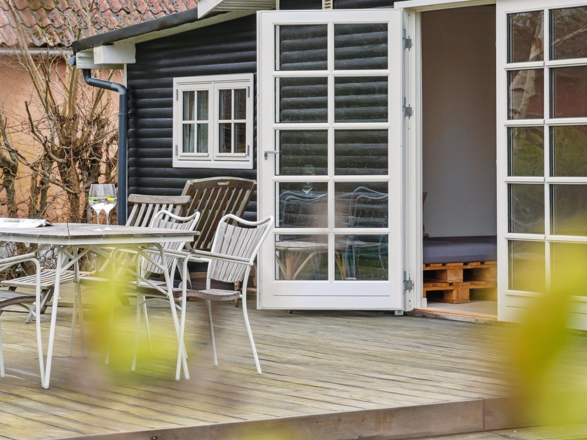 Eine Holzterrasse mit Gartenmöbeln, eine Glastür, die zu einem Raum führt, und ein Fenster mit Rollläden.