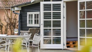 Een houten terras met buitenmeubilair, een glazen deur die naar een ruimte leidt, en een raam met luiken.