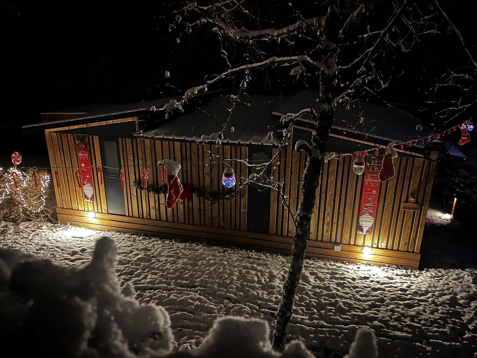 A wooden cabin with a snow-covered roof, illuminated windows, and Christmas decorations.