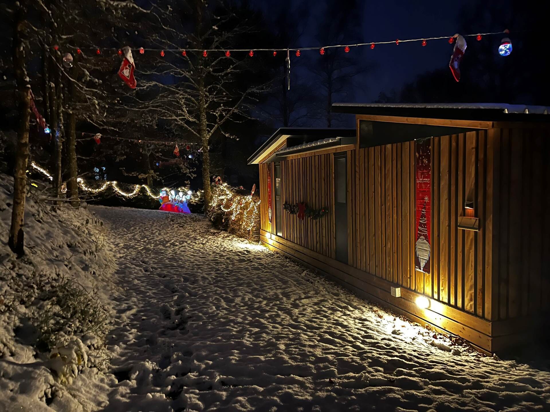 A snow-covered path leading to a wooden cabin with a 'Welcome' sign, surrounded by trees and string lights.