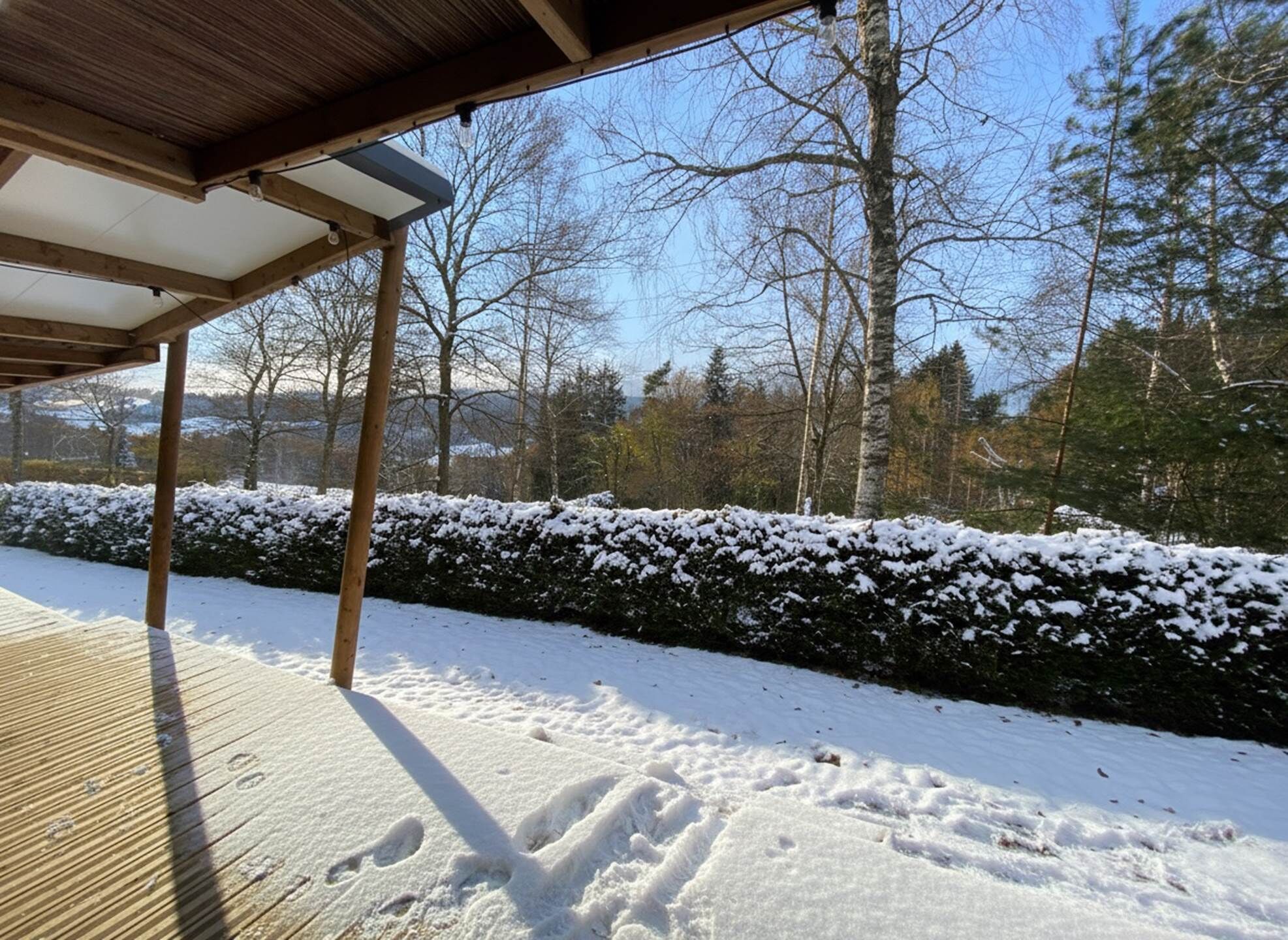 Una terraza de madera con suelo cubierto de nieve y un seto al fondo.