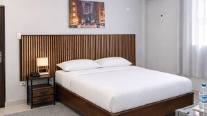 A modern hotel room with a wooden headboard, a bed with white linens, a nightstand with a lamp, a grey rug, and a small table with a bottle. - Standard Queen Room at Tris Estates Hotel & Suites (Duazon)