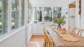 A bright kitchen with white cabinets, a wooden dining table, and a view of trees through large windows.