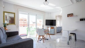 Uma sala moderna com um sofá cinzento, uma mesa de jantar com cadeiras, uma televisão e uma varanda com vista para edifícios.
