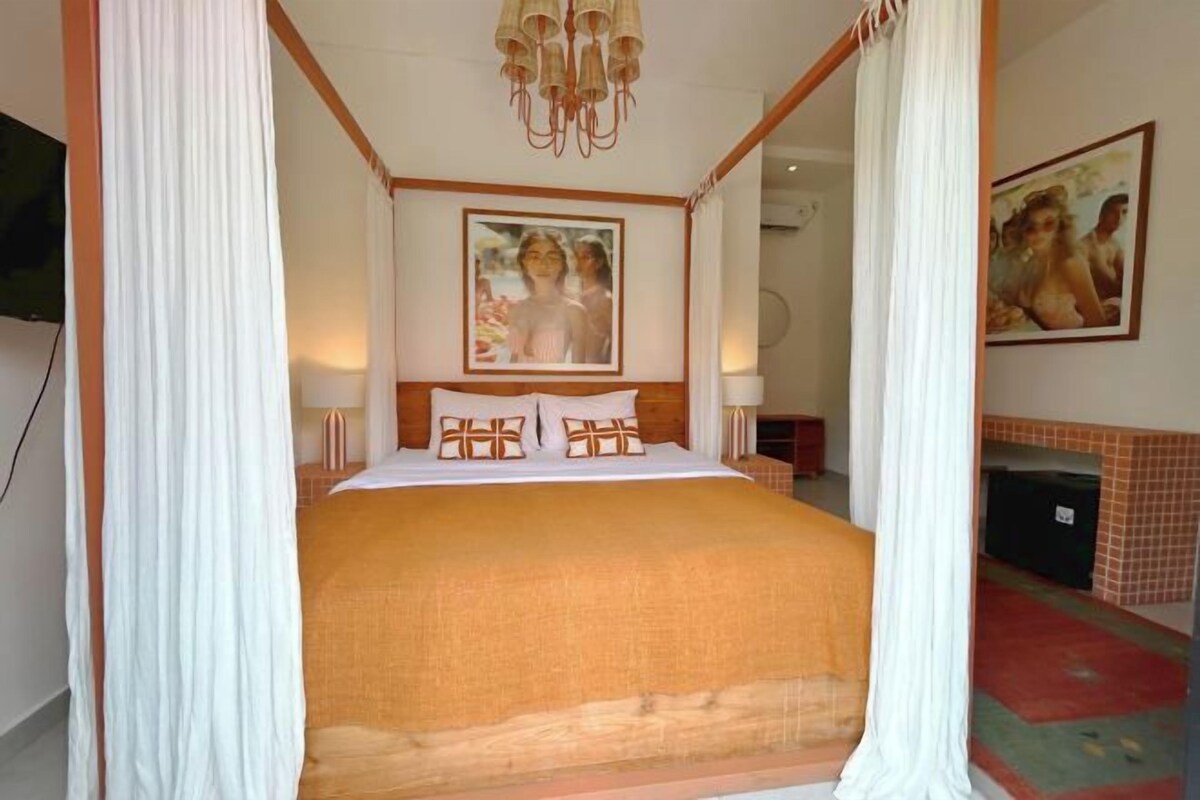 Ein Schlafzimmer mit einem Himmelbett, weißen Vorhängen, einer Chandelierleuchtre und zwei Bildern an der Wand.