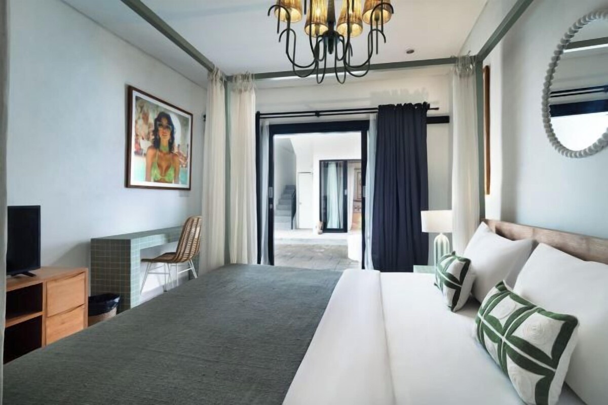 A hotel room with a large bed, a television, a chair, a painting, and a view through a glass door.