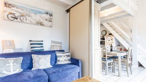 Un salon avec un canapé bleu, une table basse sur laquelle sont posées des brochures, un tableau représentant un vélo et un escalier menant à une salle à manger.