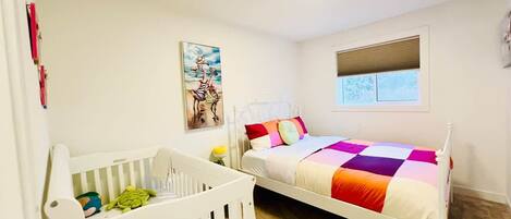 Habitación infantil con una cuna, una cama con colcha colorida, una pintura en la pared y una ventana con persianas.