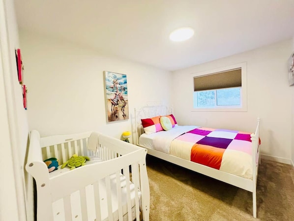 A child's room with a crib, a bed with a colorful quilt, a wall painting, and a window with blinds.