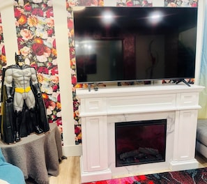 A room with a white fireplace, a flat-screen TV mounted above it, and a Batman action figure on a small table.