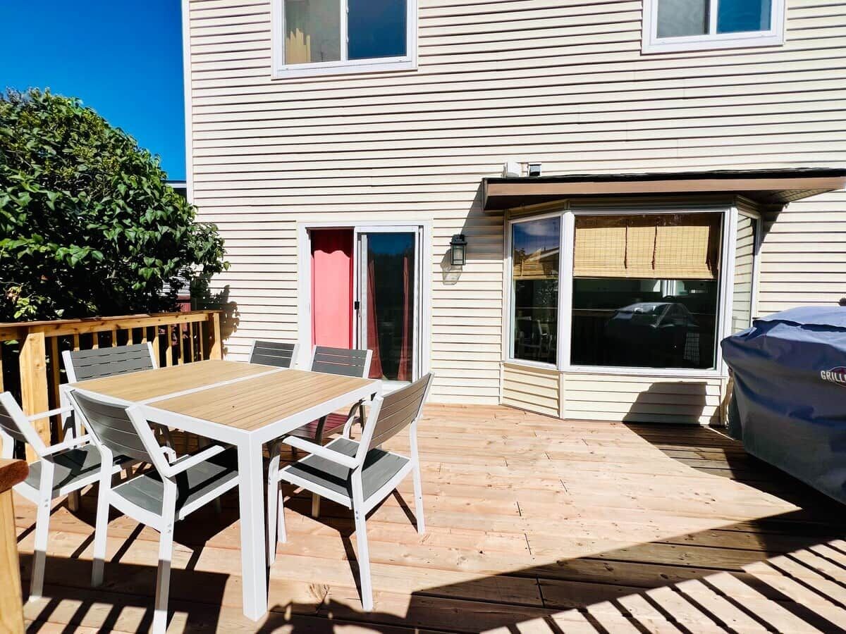 Eine Terrasse mit Holztisch und -stühlen, einem überdachten Grill und einer Glastür, die zu einer Garage führt.