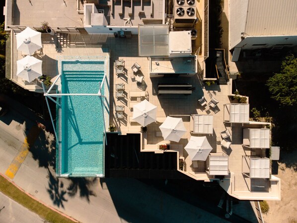 Vue aérienne d’un espace piscine sur le toit, avec plusieurs chaises longues et parasols, le tout entouré d’immeubles.