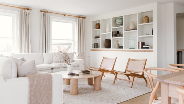 Ein modernes Wohnzimmer mit einem weißen Sofa, zwei Holzstühlen, einem runden Couchtisch und einem Bücherregal mit verschiedenen Dekorationsobjekten.