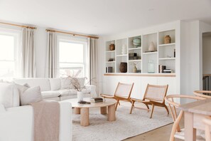 Un salon moderne avec un canapé blanc, deux fauteuils en bois, une table basse ronde et une bibliothèque agrémentée de divers objets décoratifs.