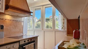 A kitchen with a stove, oven, and a window with a view of houses. - Apto 3 Dormitorios Somo - Peñasblancas 30801 (Somo)
