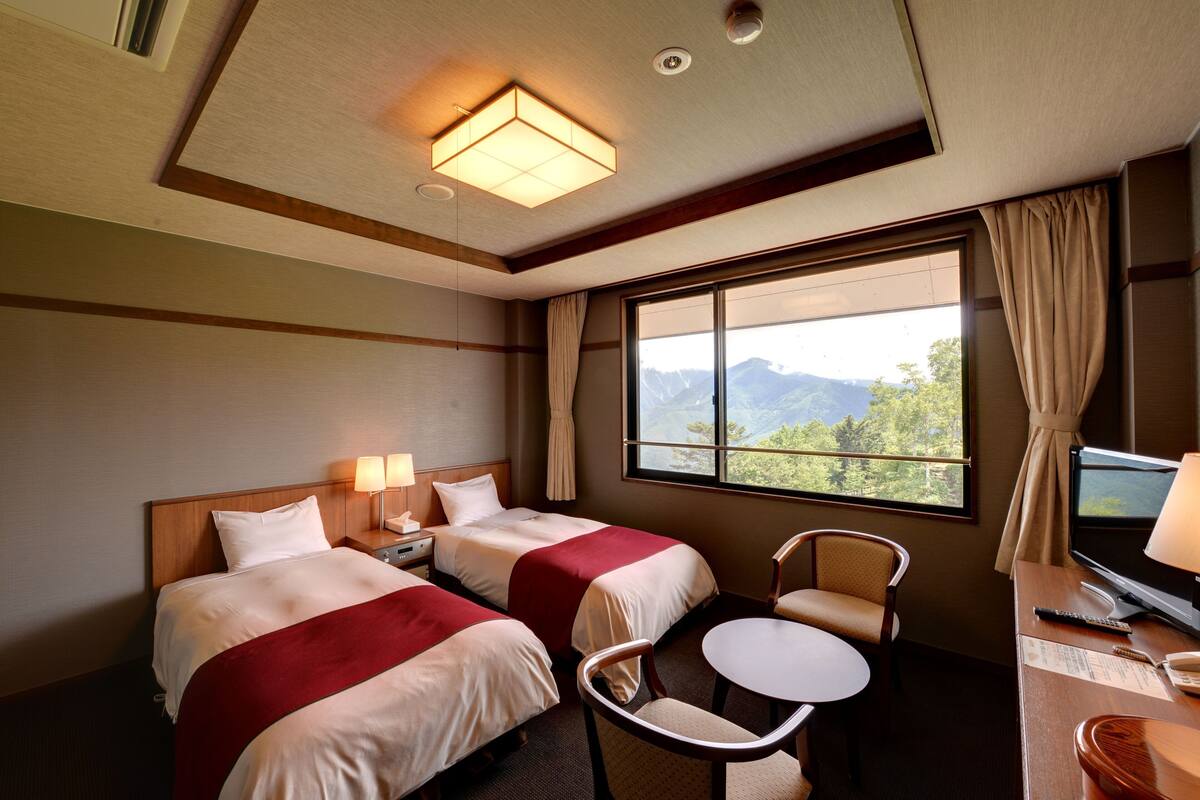 A hotel room with two beds, a small table, a chair, a TV, and a window with a mountain view.