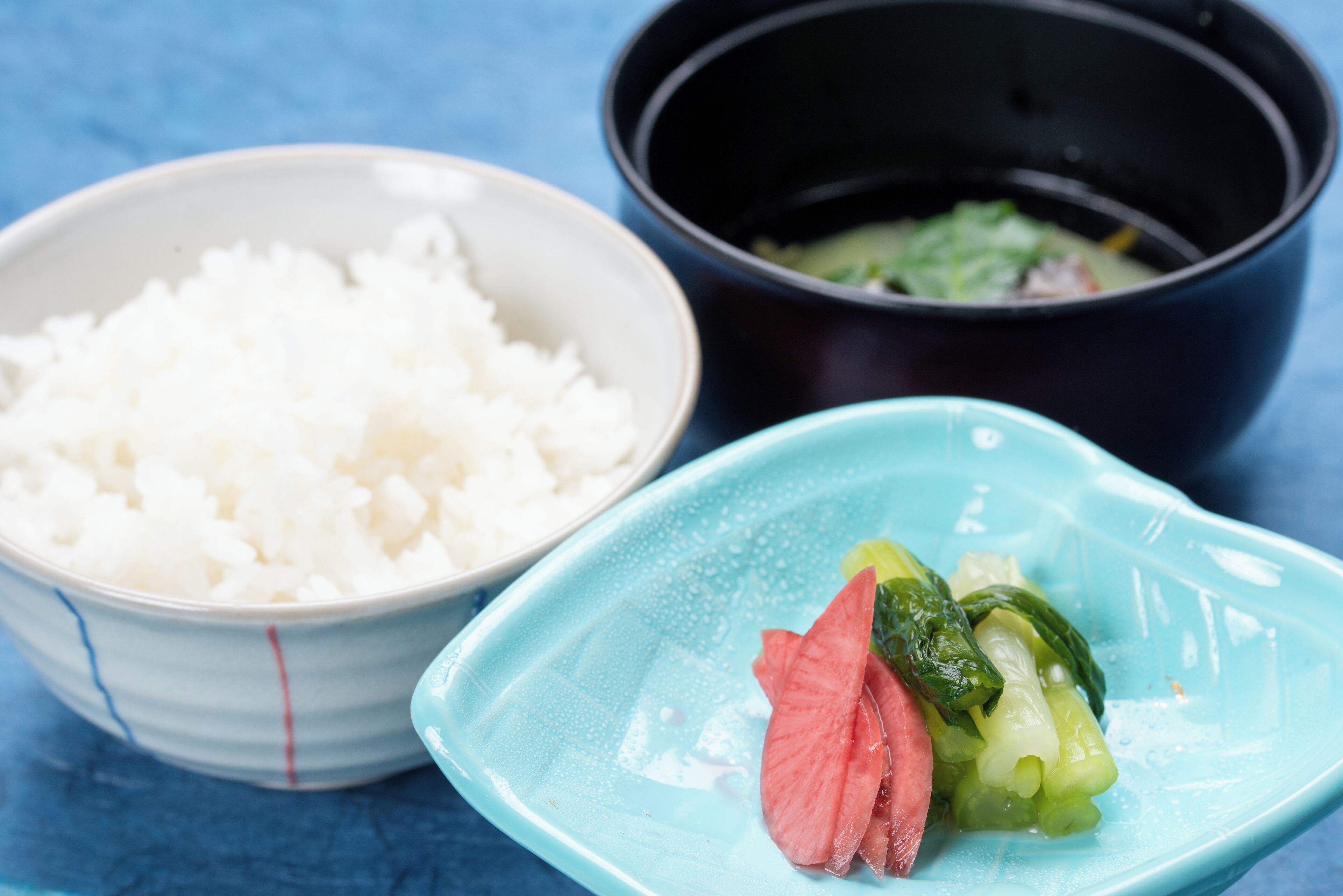 A bowl of white rice, a black bowl with a green vegetable, and a blue plate with a pink slice of meat and green vegetables.