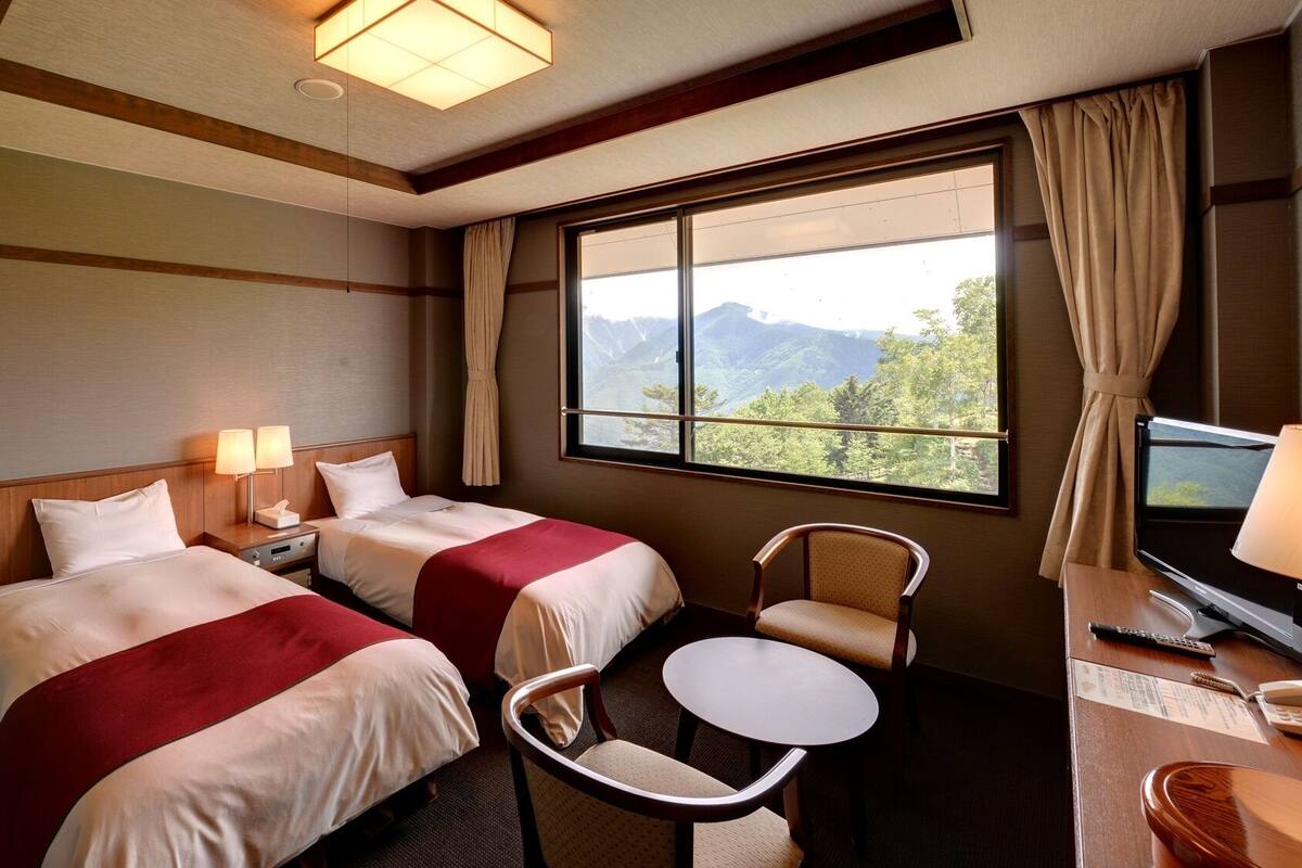 A hotel room with two beds, a desk with a chair, a TV, and a window with a mountain view.