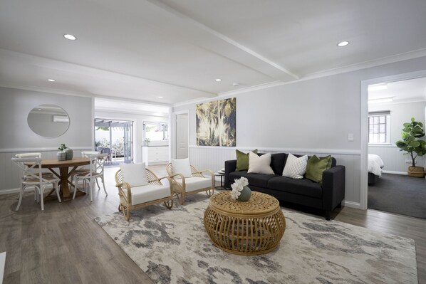 Un salon moderne comprenant un coin repas, un canapé noir agrémenté de coussins verts et blancs, des meubles en osier et un grand tableau abstrait accroché au mur.