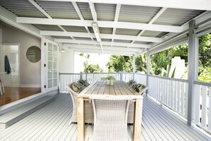 Une terrasse extérieure couverte, dotée d’une table en bois et de chaises en osier, entourée de rampes blanches et offrant une vue sur la verdure.