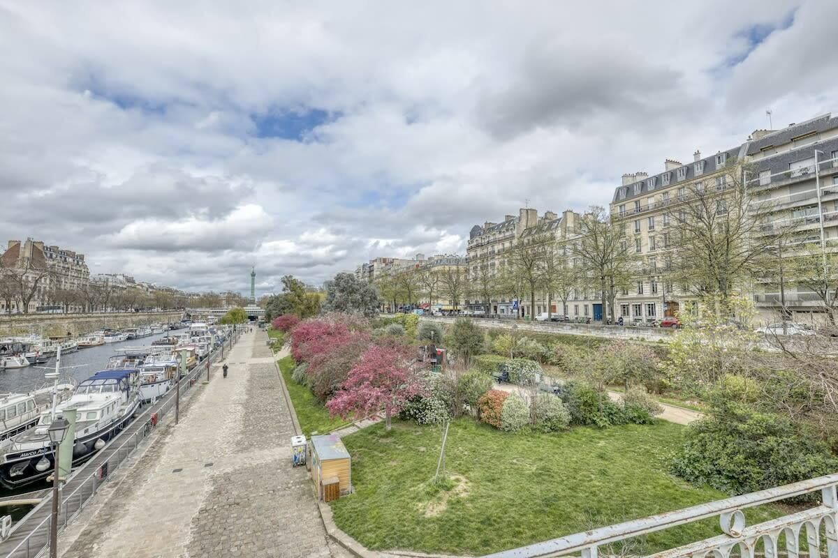 Une promenade au bord de la rivière, avec des bateaux amarrés sur la rive gauche, une zone herbeuse avec des arbres sur la rive droite et des bâtiments en arrière-plan.
