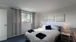 Une chambre à coucher moderne avec un grand lit, une table de chevet avec une lampe et une vue sur les arbres par la fenêtre.