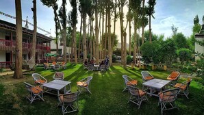 An outdoor seating area with tables and chairs, surrounded by trees and a building with a balcony. - Hotel Shambhala Resort (Leh)