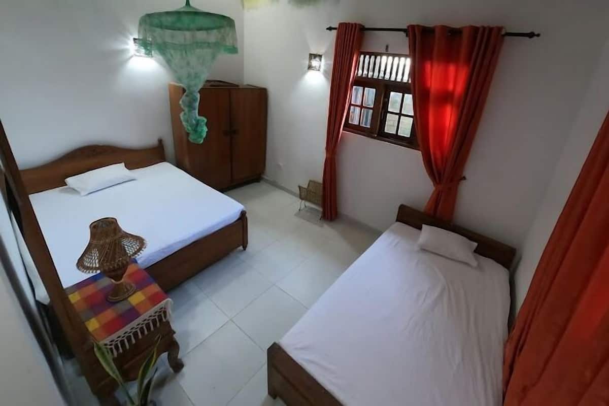 A room with two beds, a wooden wardrobe, a small table with a colorful mat, a hanging green canopy, and a window with red curtains.