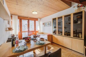 A dining area with a wooden table set for two, a kitchen with wooden cabinets, and a large window with curtains. - Slalom 73 (Bardonecchia)