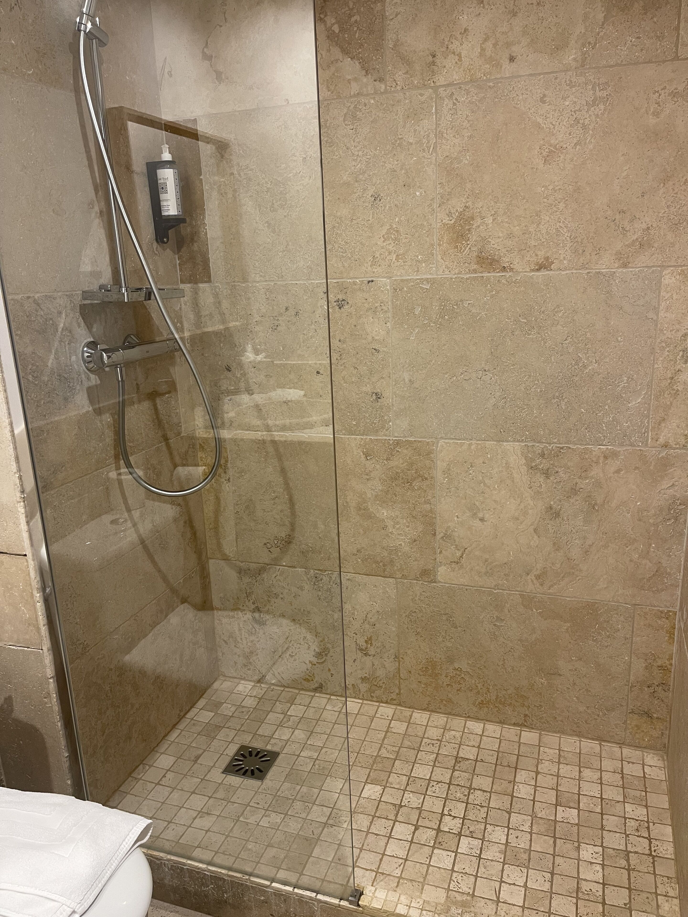 Ducha con mampara de vidrio, cabezal de ducha de mano, dispensador de jabón manual y un estante pequeño.