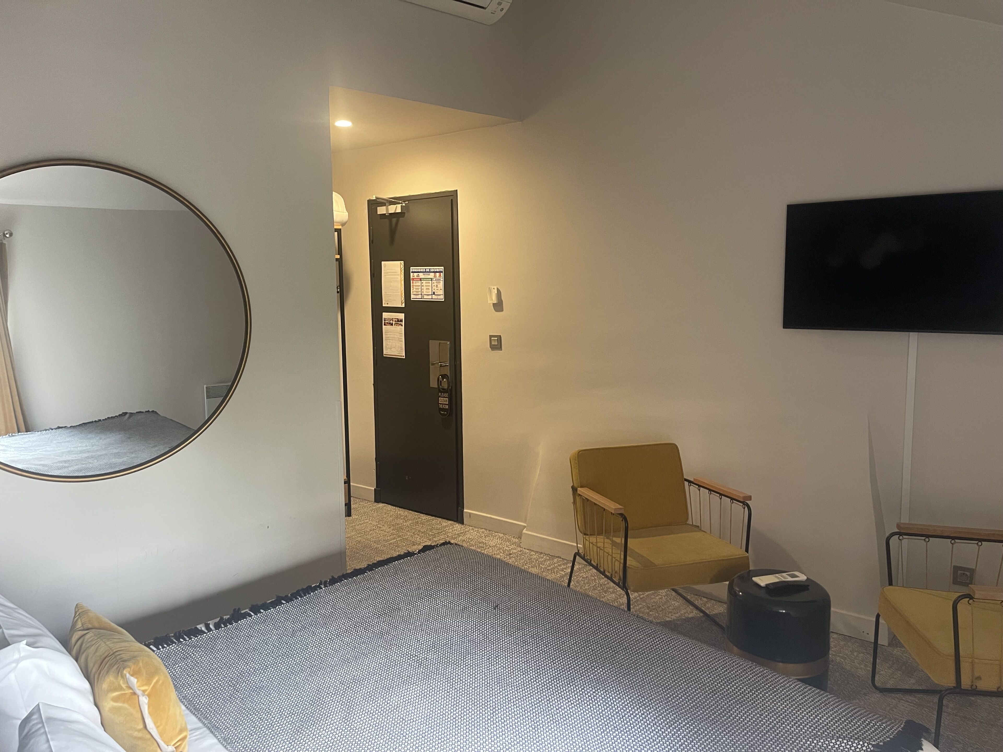 Ein Hotelzimmer mit einem Bett, einem Sessel, einem kleinen Tisch, einem Papierkorb, einem Spiegel, einer Tür mit einem Schild und einem an der Wand befestigten Fernseher.