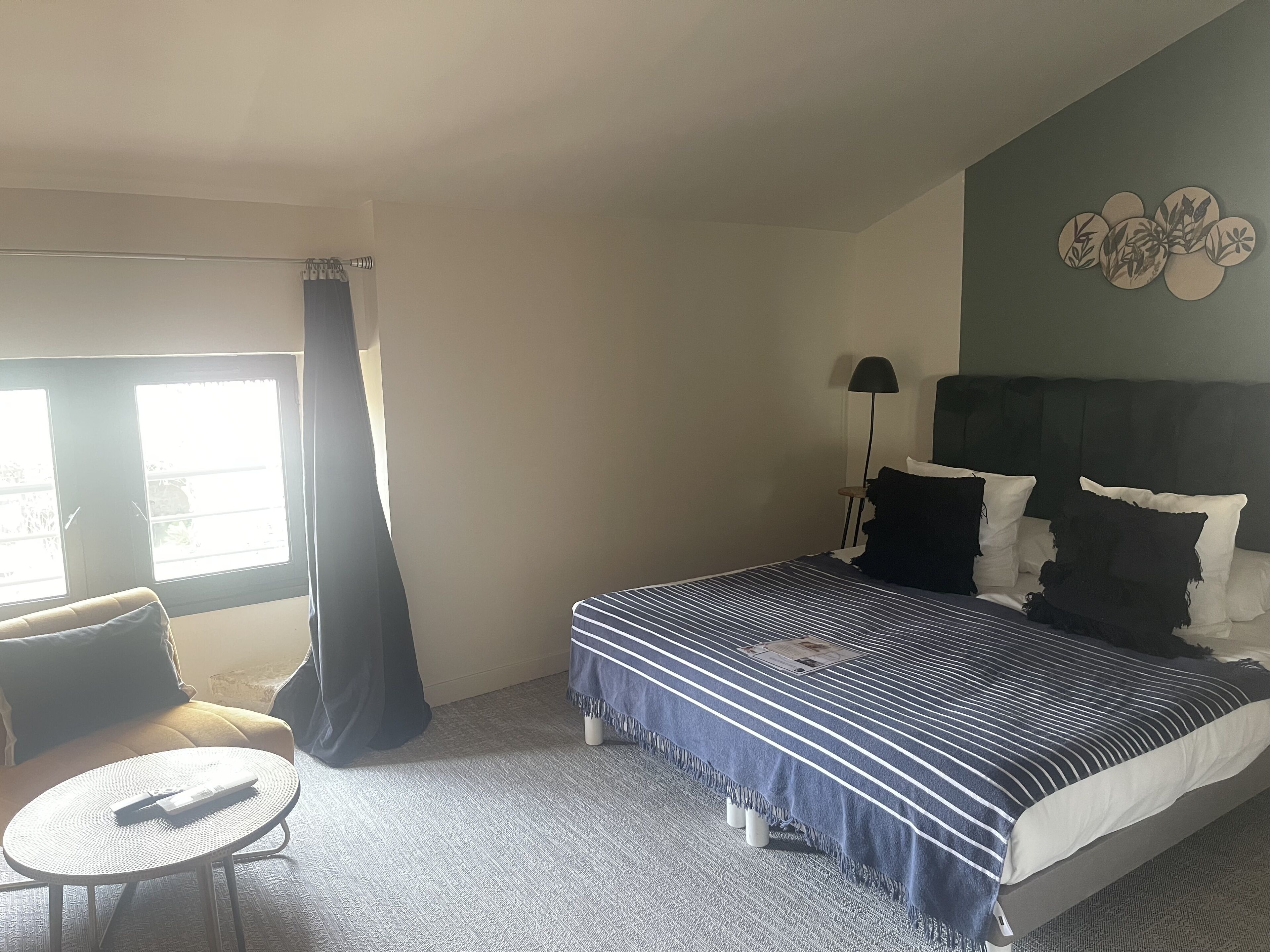 A hotel room with a bed, a small round table, a chair, a window with curtains, and a lamp.