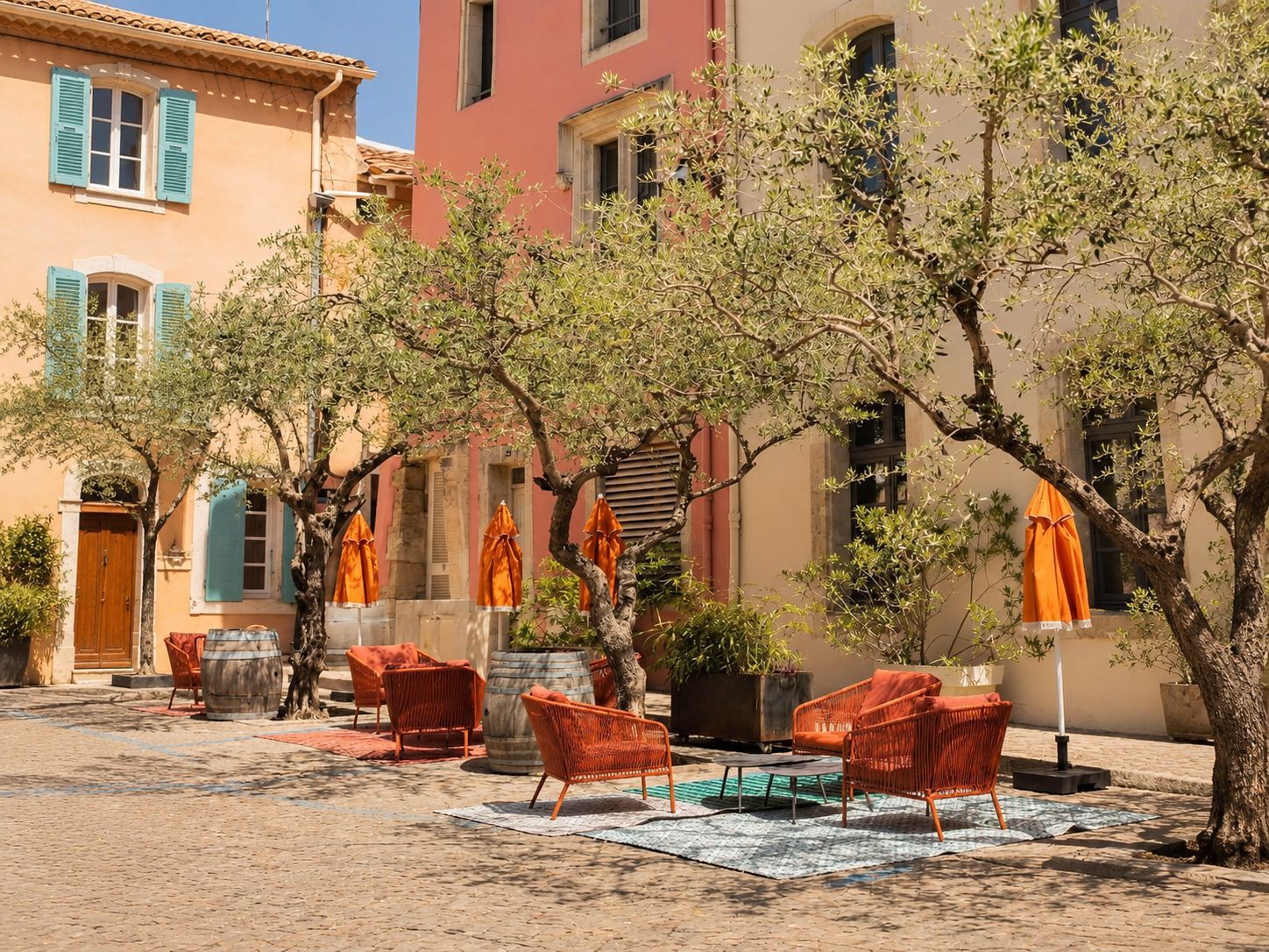 Un área de estar al aire libre con sillas de mimbre y sombrillas naranjas, rodeada de árboles y edificios con fachadas de colores pastel.