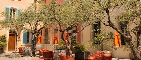 Un área de estar al aire libre con sillas de mimbre y sombrillas naranjas, rodeada de árboles y edificios con fachadas de colores pastel.
