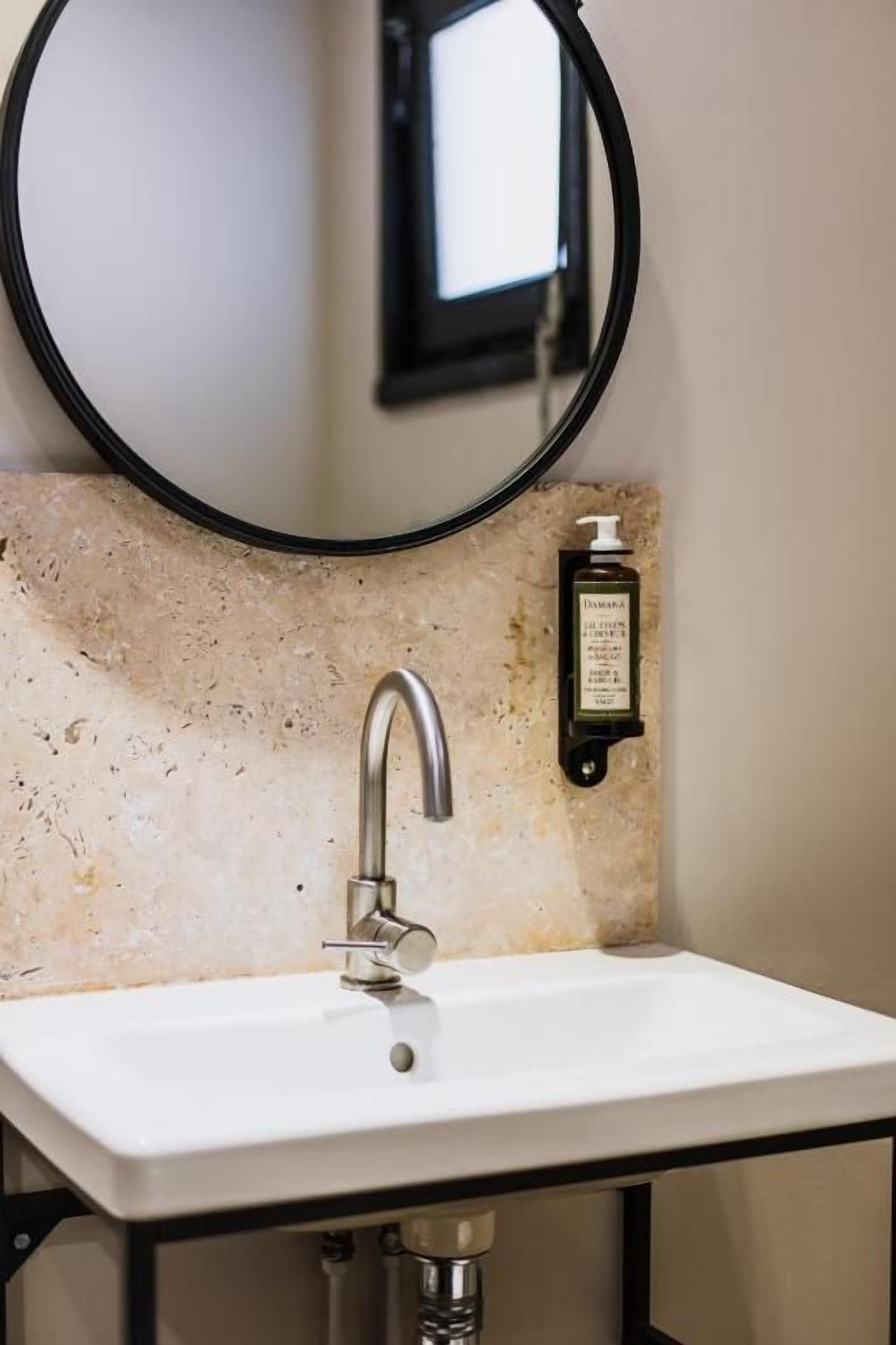 A bathroom sink with a mirror above it, a soap dispenser, and a wall-mounted light.