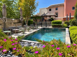 Una piscina con sillones de descanso y sombrillas, rodeada de vegetación y un muro de piedra.