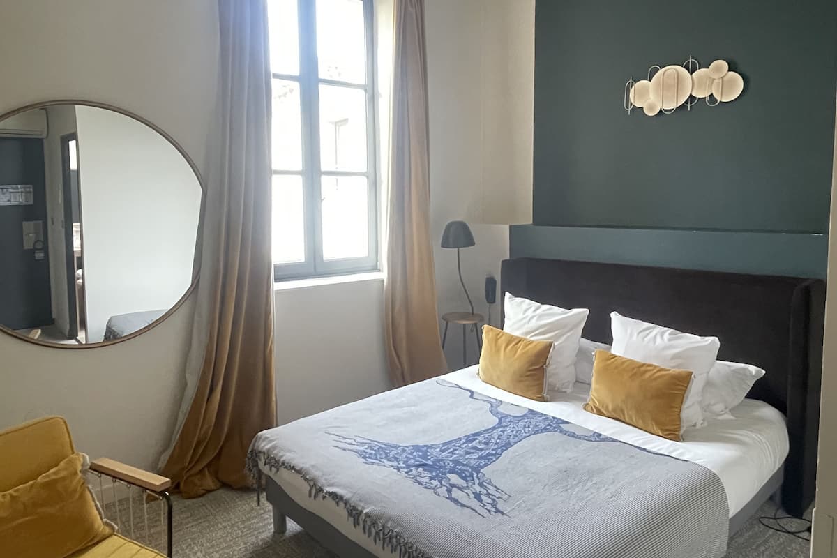 A hotel room with a bed, a round mirror, a window with curtains, a lamp, and a wall-mounted light fixture.