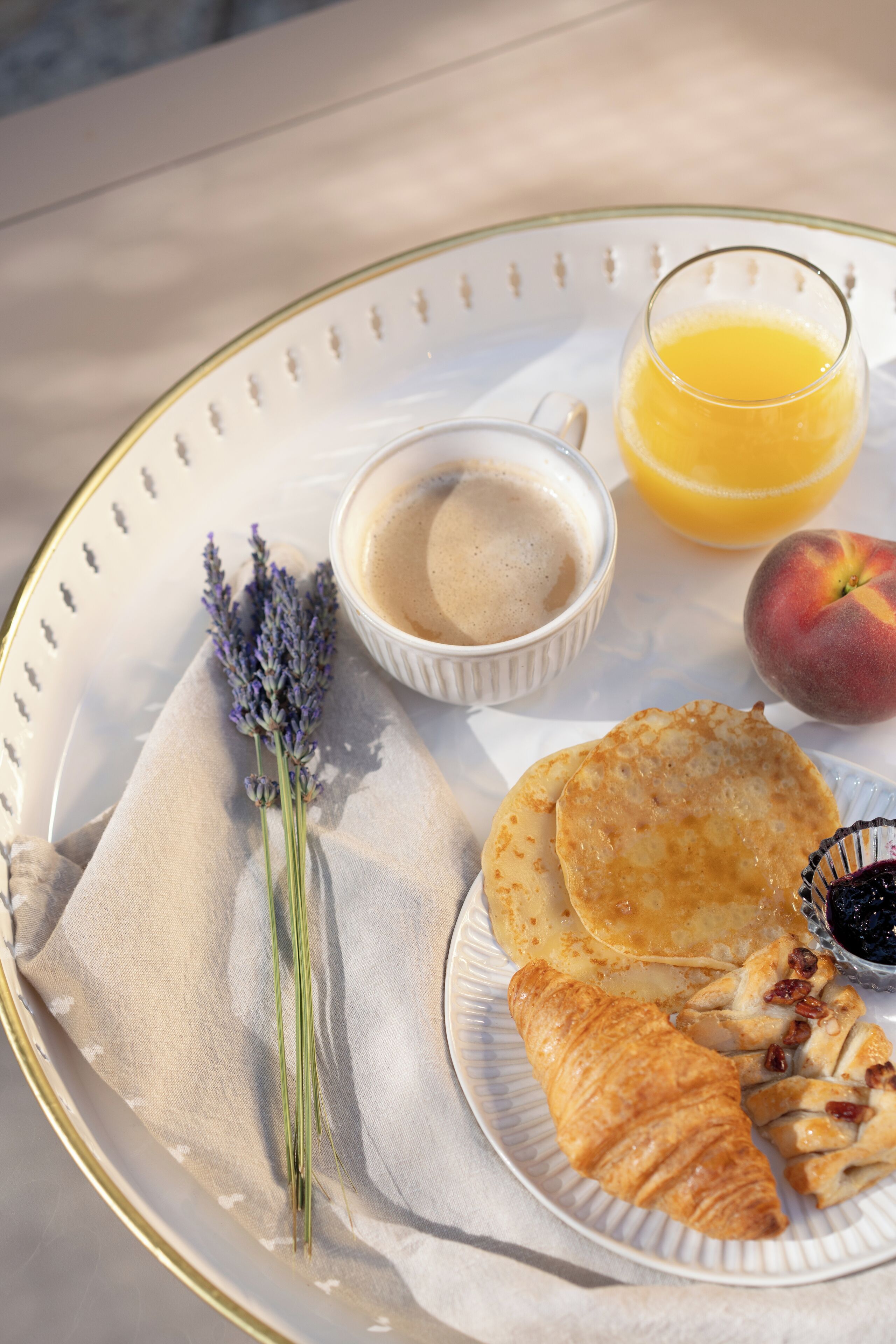 Una bandeja con panqueques, un croissant, un melocotón, un vaso de jugo de naranja, una taza de café y una ramita de lavanda.