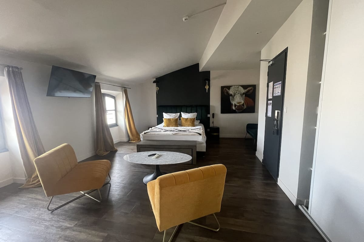 A modern hotel room with a bed, two yellow armchairs, a round table, and a large painting on the wall.