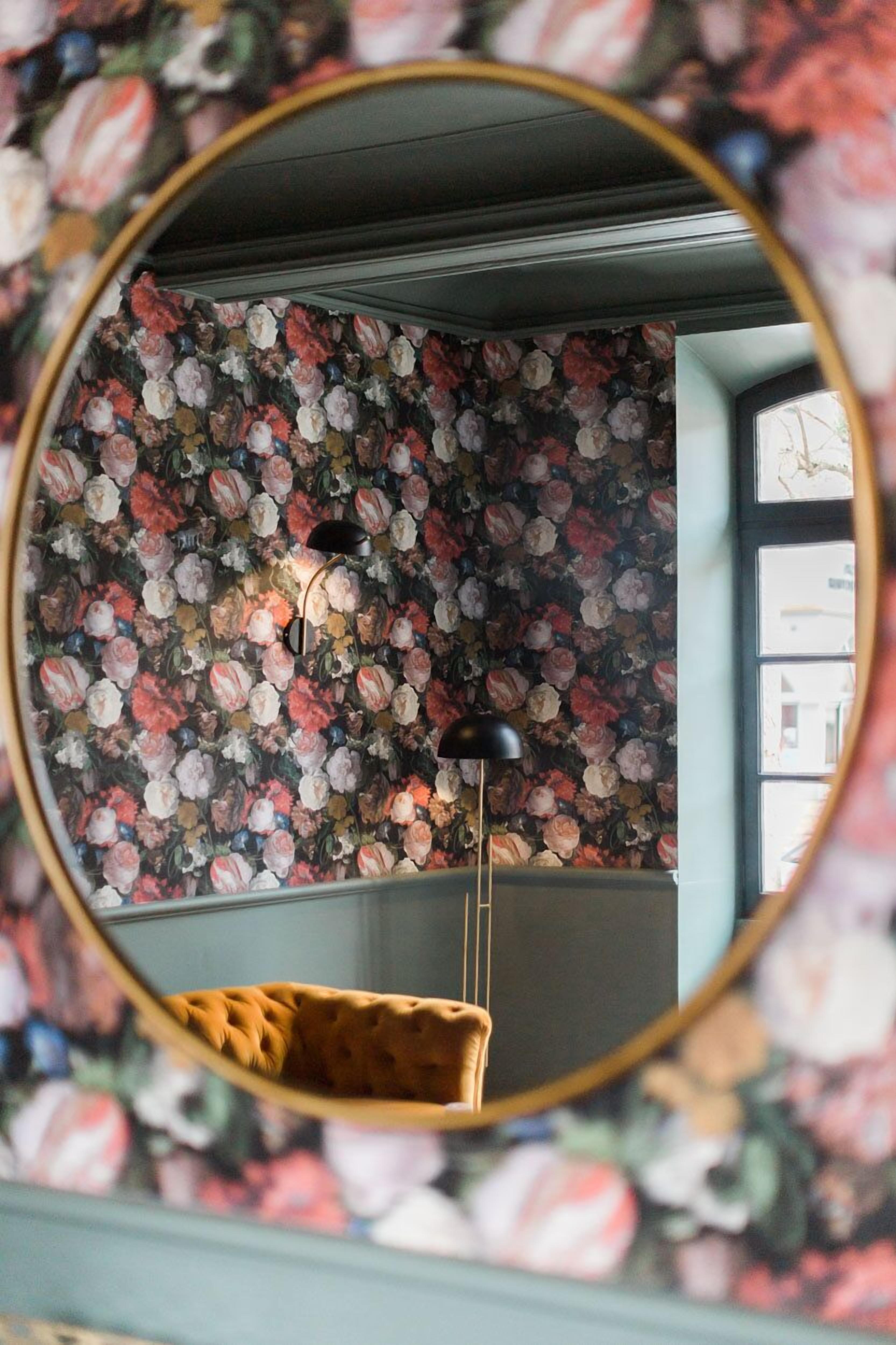 Ein Zimmer mit floralem Wandpapier, einem runden Spiegel und einer hängenden Leuchte.