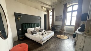 A bedroom with a green upholstered headboard, a white bed with patterned pillows, a red ottoman, a small desk, and a marble side table. - Victoria Boutique Hôtel (Saint-Paul-Trois-Chateaux)