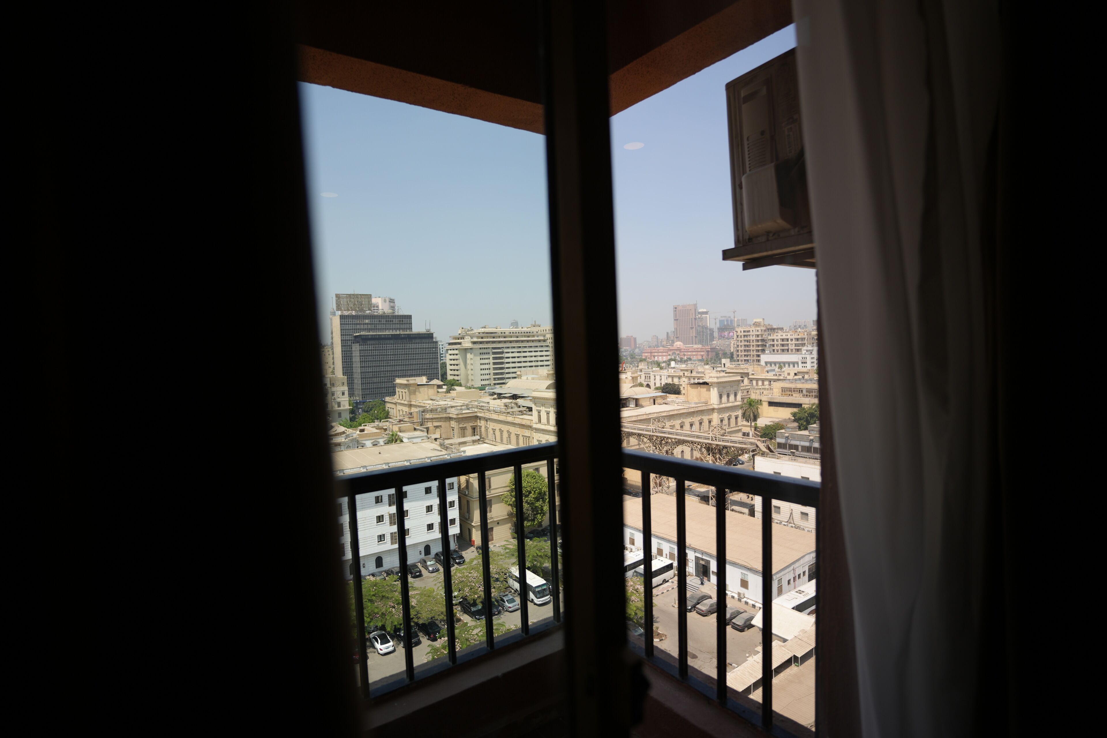 A view from a window with a metal railing, overlooking a cityscape with various buildings and a clear sky.