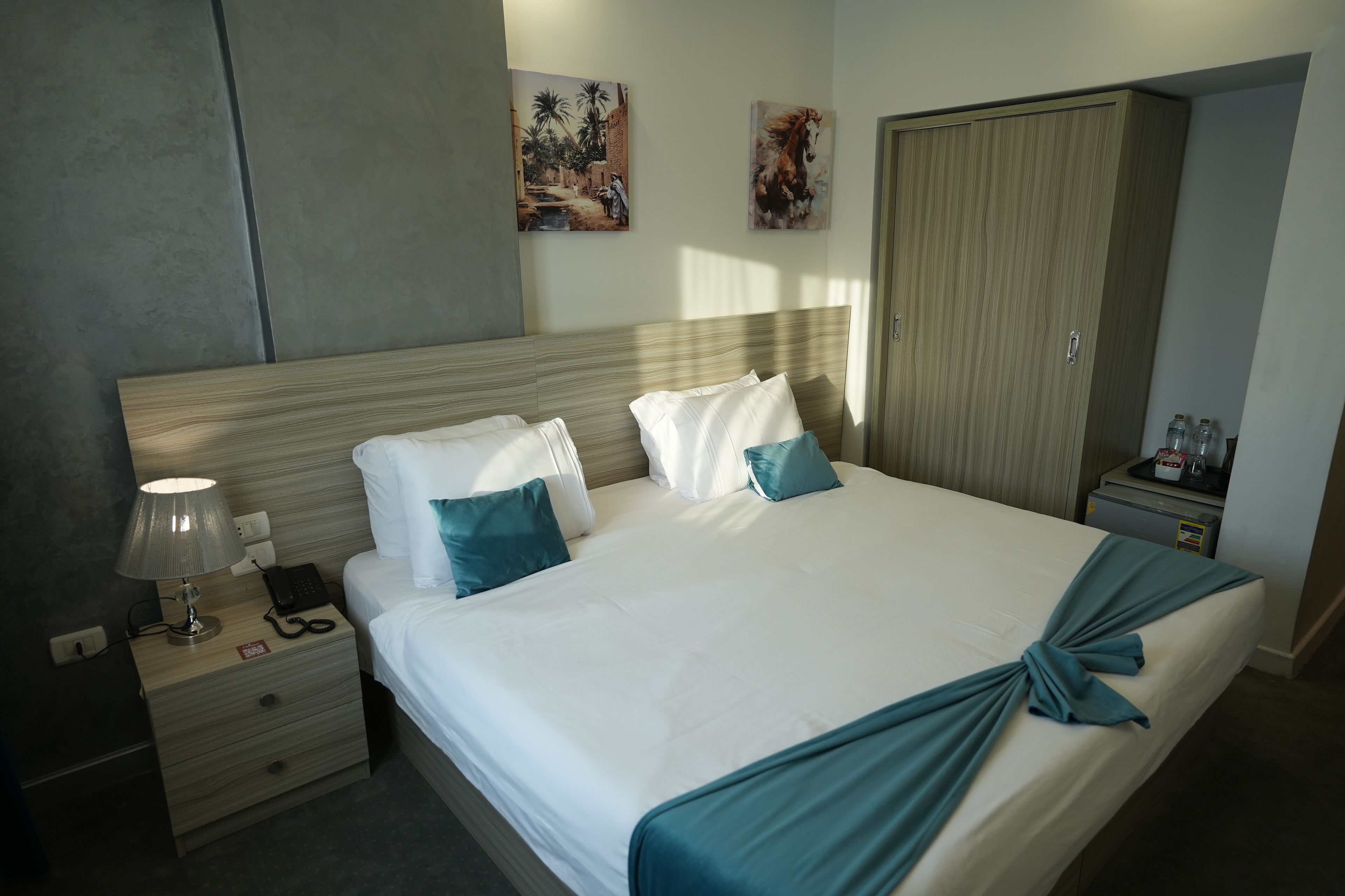 A hotel room with a large bed, two pillows, a bedside table with a lamp, a phone, and a nightstand with a drawer. There is a wardrobe and a wall with two paintings.