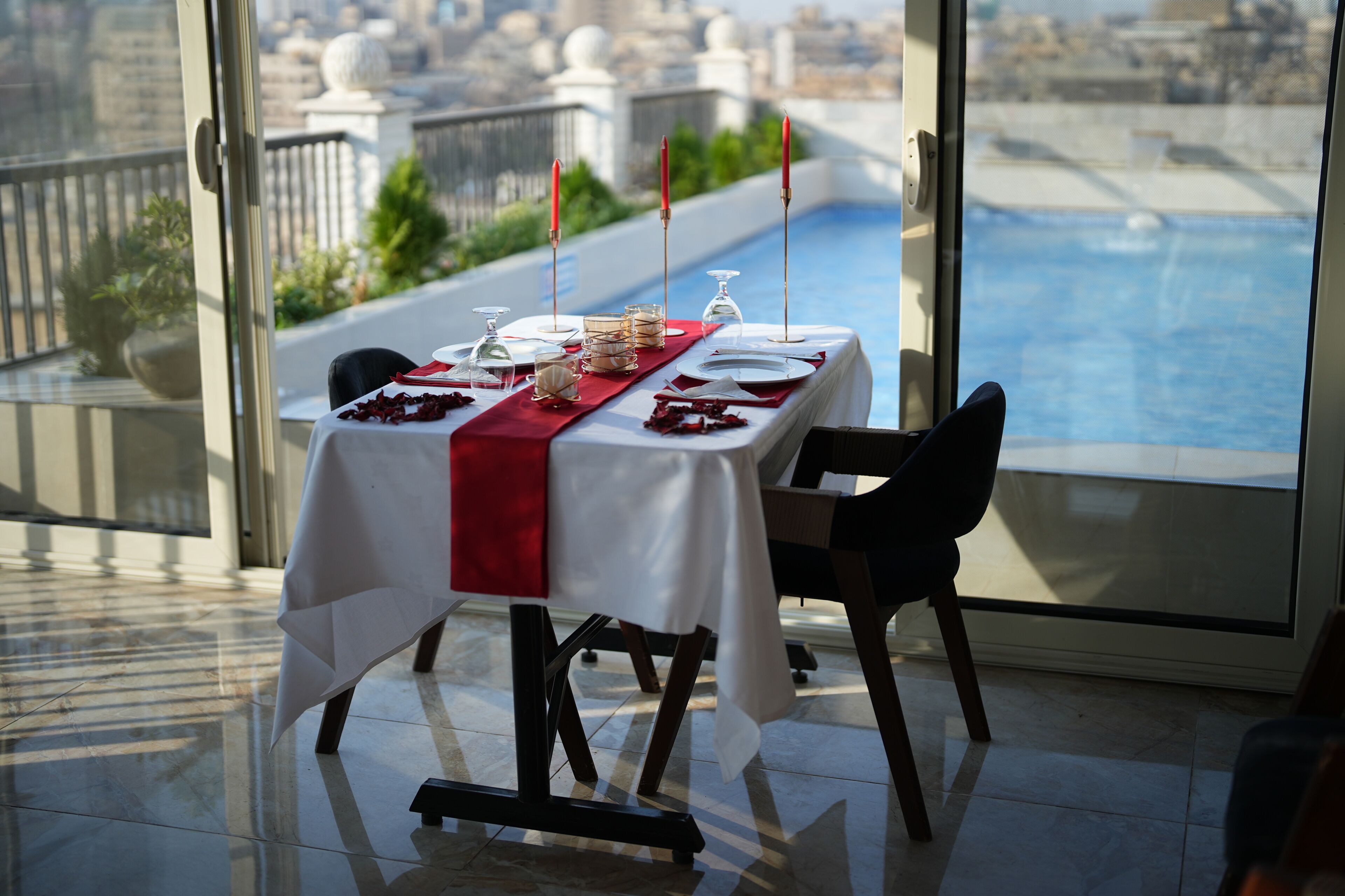 Une table à manger dressée pour deux personnes, avec un chemin de table rouge, des verres et des couverts, donnant sur une piscine et un paysage urbain.