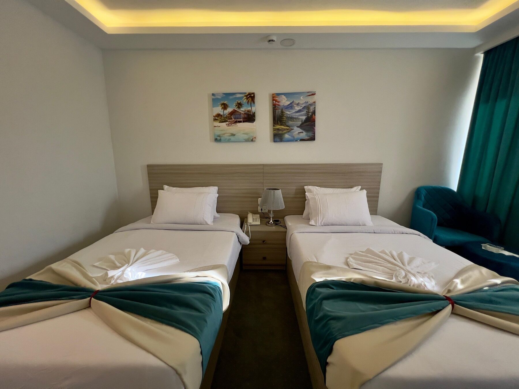 Una habitación de hotel con dos camas, una mesita de noche, una lámpara y dos cuadros en la pared.